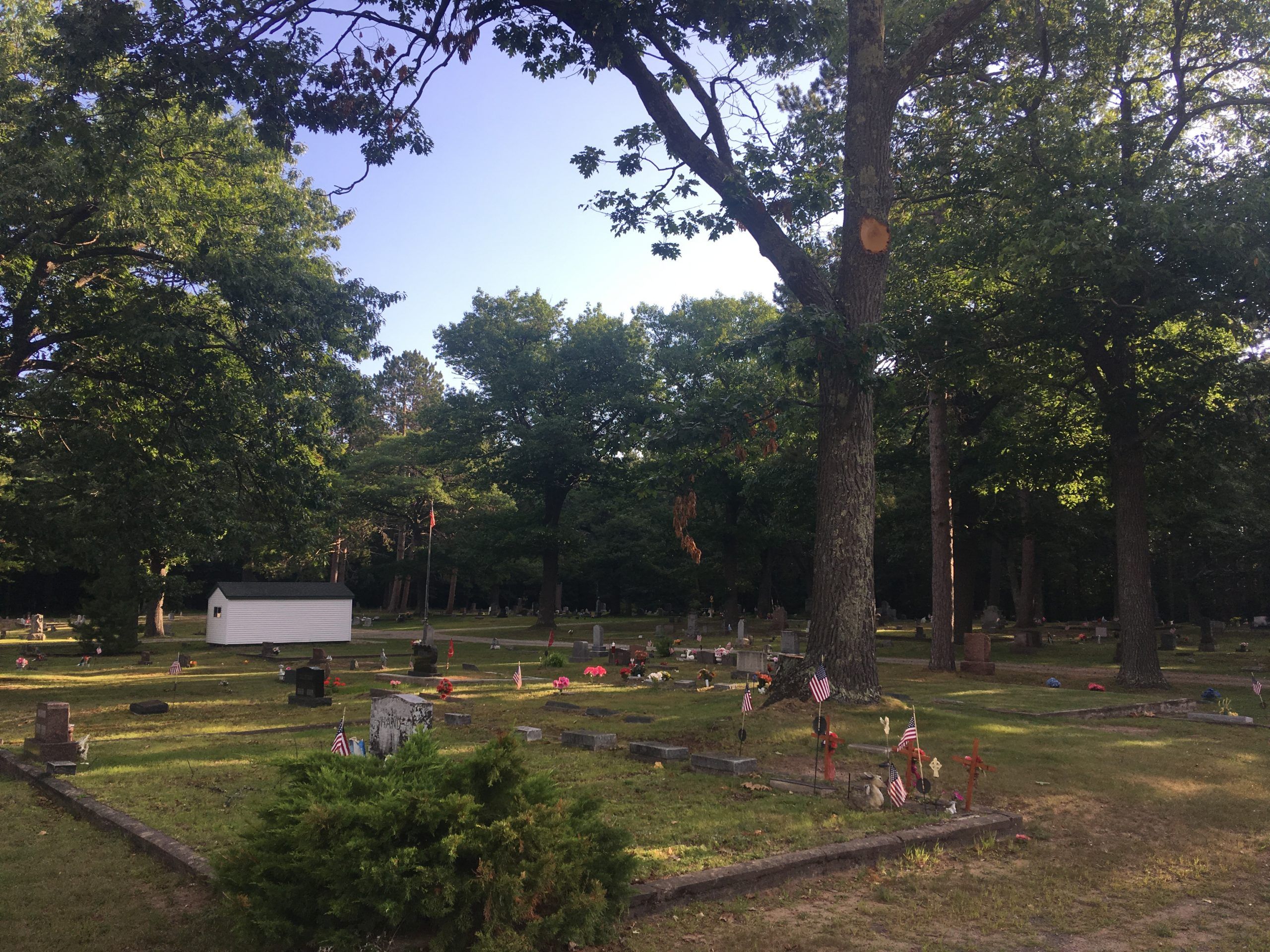 View of cemetery headstones with flowers and decorations. Trees are also throughout the cemetery. And a little white building.